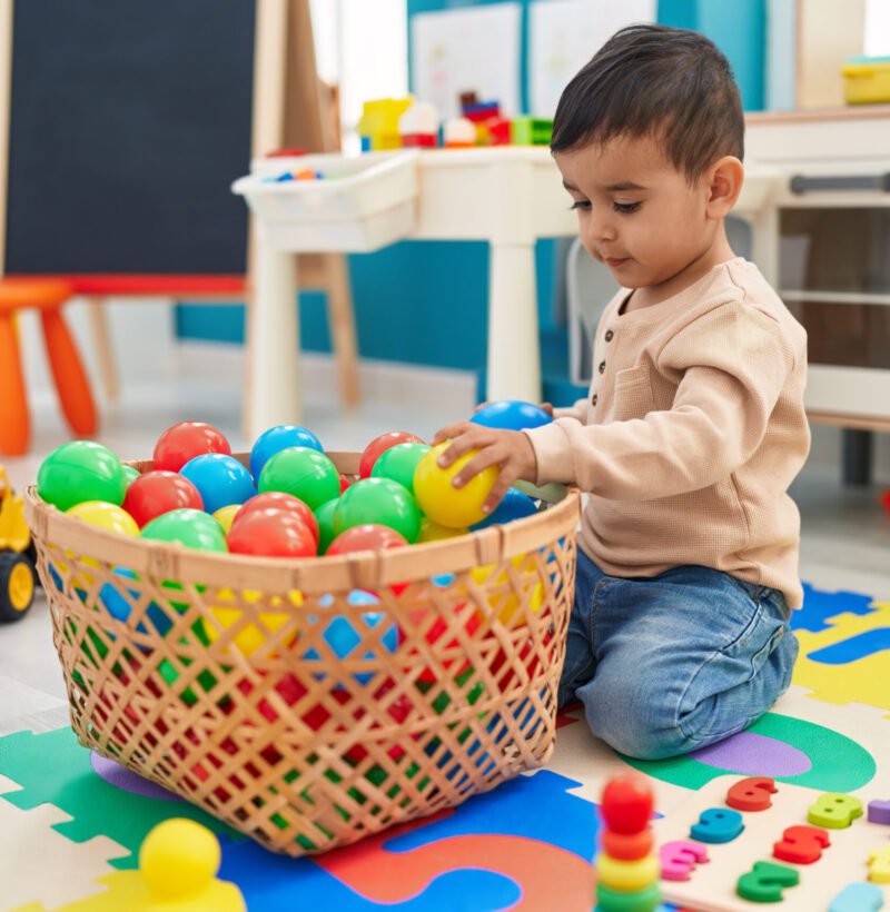 Adorable hispanic toddler playing with balls sitting on floor at kindergarten